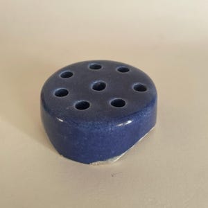May include: A blue ceramic flower frog with nine holes. The flower arranging tool is a rounded shape with a flat bottom and a glossy finish. The ceramic has a slightly textured appearance.