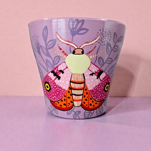 May include: A hand-painted, lilac-coloured ceramic pot with a colourful moth design. The moth has pink, orange, and black details, with a cream-coloured head and feathery antennae. The pot is decorated with floral and leaf patterns.