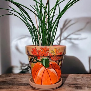 May include: A decorative ceramic pumpkin planter with a matching saucer, holding a green plant. The planter is painted with a large orange pumpkin and autumn leaves. The planter sits on a wooden surface.