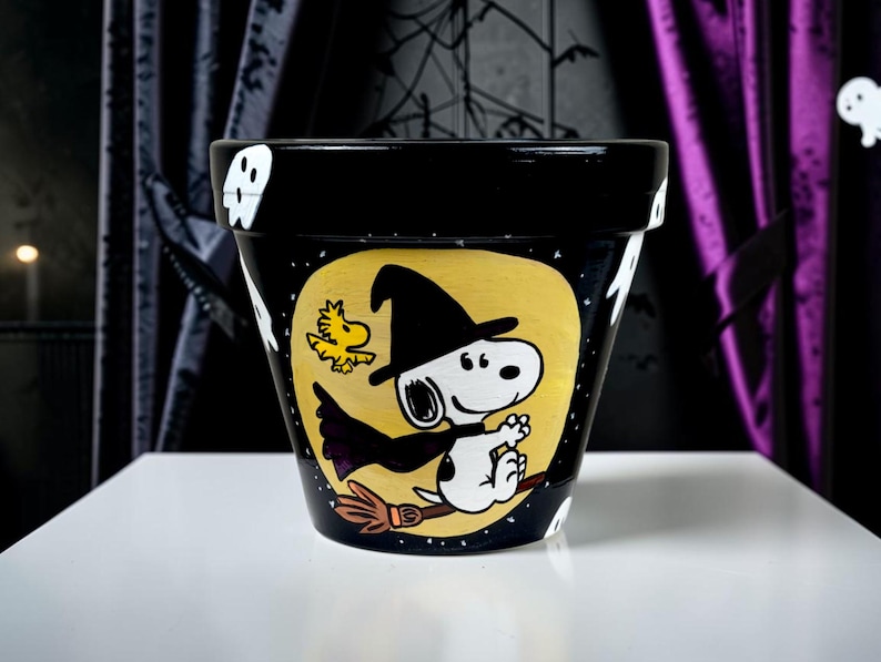 以下が含まれることがあります： 黒い陶器製の植木鉢。黒い魔女の帽子とマントを身につけ、黄色い月を背景にほうきに乗ったスヌーピーと、小さな黄色い鳥が手描きで描かれている。鉢の縁には白いお化けの絵が施されている。