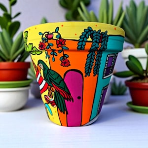 May include: A brightly painted ceramic plant pot with a colourful design. The pot features a parrot, a door, and a window, set against yellow, orange, and teal. Red flowers and green foliage add detail to this decorative planter, perfect for indoor plants.
