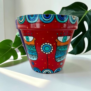 Peut inclure: Pot de fleur en terre cuite rouge peint à la main avec un design fantaisiste. Le pot présente des figures stylisées bleues et sarcelle en forme d'yeux et un motif floral. Le bord est décoré de motifs bleus et sarcelle.