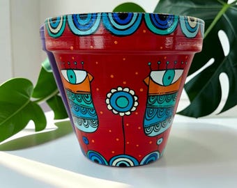以下が含まれることがあります： 手描きの赤いテラコッタ植木鉢。青とターコイズブルーの目のような図形と花のモチーフが特徴です。縁は青とターコイズブルーの模様で装飾されています。ユニークなデザインの植木鉢です。