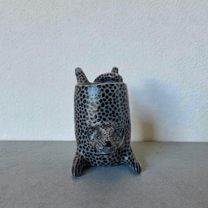 Peut inclure: Figurine de léopard en céramique noire et blanche. Le léopard est représenté dans une pose ludique, avec ses pattes et sa tête tendues vers le bas. Le corps est recouvert de petits points noirs et le visage présente des détails sculptés.