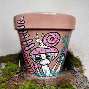 May include: A terracotta planter painted with a pink and white mushroom, a snail, and green foliage. The planter is a light brown color and has a smooth, matte finish.
