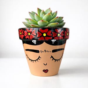 May include: A terracotta flower pot with a painted face design, featuring closed eyes, a red lip, and a black floral border. The pot holds a green succulent plant. The pot's design is inspired by Frida Kahlo.
