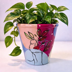 May include: A hand-painted ceramic plant pot with a face design and a vibrant green plant. The pot features a woman's face with closed eyes, red lips, and a floral design. The pot is filled with a lush green plant.