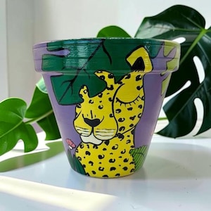 May include: A hand-painted ceramic flower pot with a whimsical design. The pot features a yellow leopard with black spots against a purple background. Green leaves and foliage are painted around the rim. The pot is suitable for indoor or outdoor use.