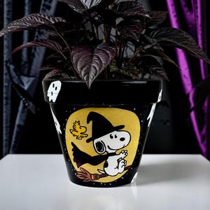 以下が含まれることがあります： 黒い陶器製の植木鉢。魔女の仮装をしてほうきに乗ったスヌーピーとウッドストックの手描きイラストが描かれています。デザインは黄色い月を背景にしており、黒い縁には小さな白いお化けが描かれています。