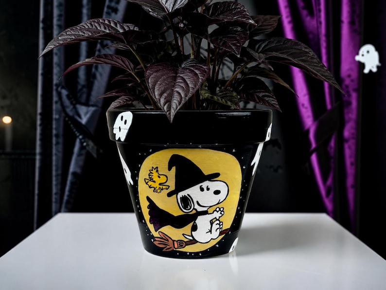 以下が含まれることがあります： 黒い陶器製の植木鉢。魔女の仮装をしてほうきに乗ったスヌーピーとウッドストックの手描きイラストが描かれています。デザインは黄色い月を背景にしており、黒い縁には小さな白いお化けが描かれています。