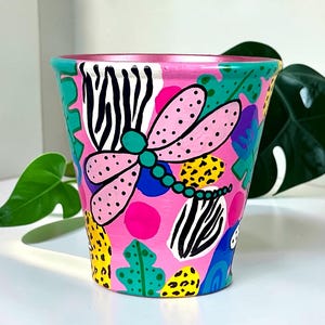 May include: A hand-painted pink ceramic plant pot with a colourful design. The pot features a dragonfly, zebra stripes, and leaf patterns in teal, yellow, and black. The rim is a metallic pink.