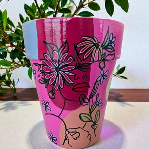 May include: A hand-painted pink plant pot with a floral design and a woman's face. The pot features a mix of pink shades, with a light blue stripe and a hand-drawn illustration of a face and flowers. Suitable for indoor or outdoor use.