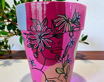 以下が含まれることがあります： 手描きのピンク色の植木鉢。花柄と女性の顔が描かれています。ピンク色の濃淡、水色のストライプ、手描きの顔と花のイラストが特徴です。屋内または屋外での使用に適しています。