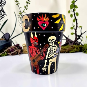 May include: A black painted flower pot with Day of the Dead artwork. The pot features a red devil skeleton playing a guitar and a white skeleton holding a book. The rim has a yellow and black design with a moon, heart, and sun.