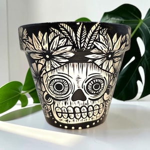 May include: Black and white ceramic plant pot with a skull design. The pot has a black base with white floral and leaf patterns around the rim and skull details on the front. The skull has large eye sockets and a detailed jawline.