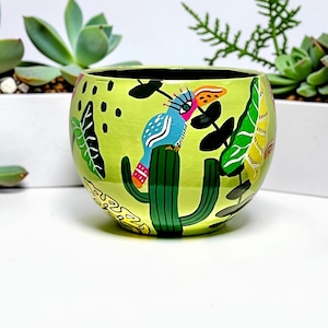 May include: A round, lime green ceramic planter featuring a hand-painted design of a blue bird with a yellow beak perched on a dark green cactus. The surface is decorated with black abstract dots and various green and yellow tropical leaf illustrations.