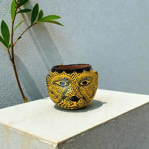May include: A yellow and black ceramic pot with a jaguar face design. The pot has a round shape with a black rim and is decorated with black dots and lines. The eyes are blue. The pot is sitting on a white surface.