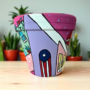 May include: Hand-painted terracotta flower pot featuring a colorful cityscape design. The pot showcases a vibrant palette of pink, purple, and white, with architectural elements and a flag motif. Ideal for indoor or outdoor plants.