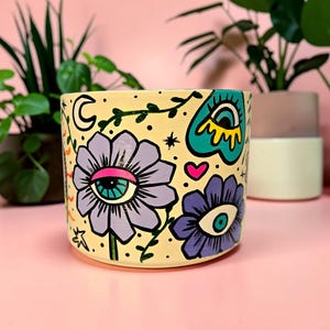 May include: A hand-painted, cylindrical ceramic planter with a cream base. The planter features floral and eye designs in purple, teal, and pink, with black accents. The design includes a crescent moon, stars, and a heart. Suitable for indoor plants.