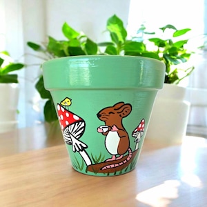 May include: A light green ceramic flower pot with a whimsical design. The pot features a brown mouse holding a cup, a red and white mushroom, and a small yellow bird. The design is set against a backdrop of green grass.