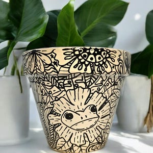 May include: A beige ceramic planter with a black and white doodle design featuring a frog inside a flower. The design includes leaves, swirls, and other abstract shapes.