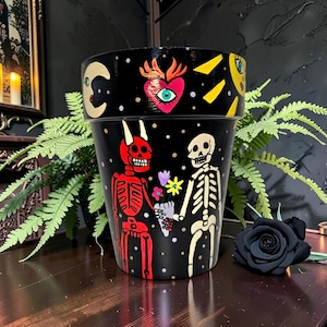 May include: Black painted flower pot with a celestial and skeleton design. The pot features a red devil skeleton and a white skeleton holding flowers. The top of the pot has a yellow sun, a crescent moon, and a heart with an eye.