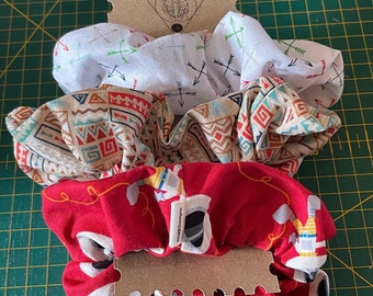 May include: Three fabric hair scrunchies stacked on a cardboard display card. The top scrunchie is white with small arrows. The middle one features a tan background with geometric patterns. The bottom red scrunchie displays a space-themed print.