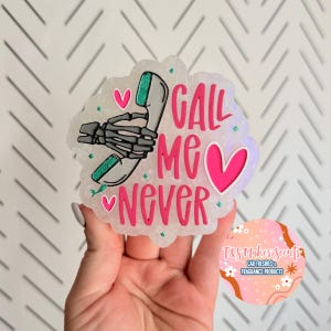May include: A car air freshener with a unique design. The air freshener features the text "CALL ME NEVER" in pink, with a skeleton hand holding a phone. The design includes pink hearts and teal accents.
