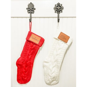 May include: Two knitted Christmas stockings with personalized leather name tags. The stocking on the left is red and the stocking on the right is white.