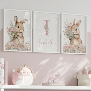 May include: Three white framed prints with a pink background. The centre print has a pink letter I with a floral design and the name Isabella. The left and right prints feature a watercolour illustration of a brown bunny holding a bouquet of flowers.