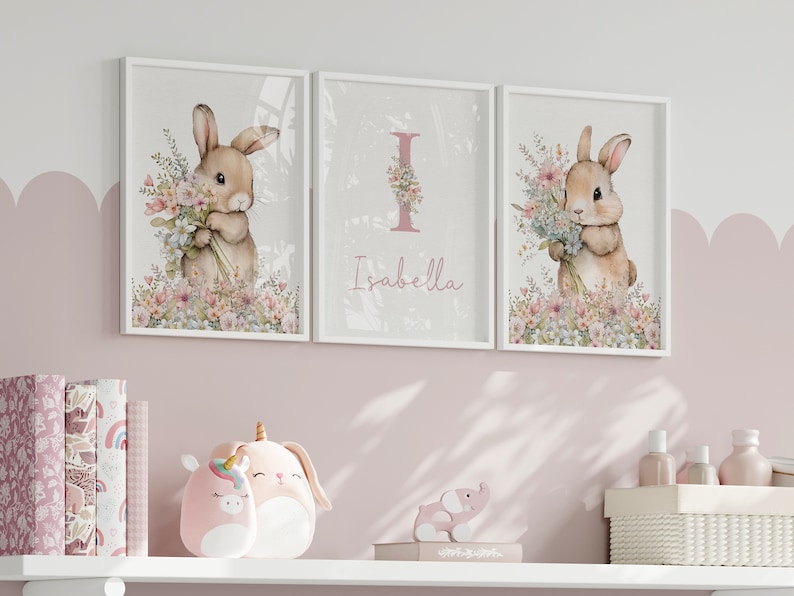 May include: Three white framed prints with a pink background. The centre print has a pink letter I with a floral design and the name Isabella. The left and right prints feature a watercolour illustration of a brown bunny holding a bouquet of flowers.