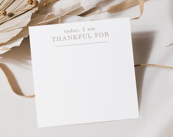 May include: A white square notepad with the text "today, I am THANKFUL FOR" in a light brown font. The notepad is placed on a white surface with dried plants in the background.