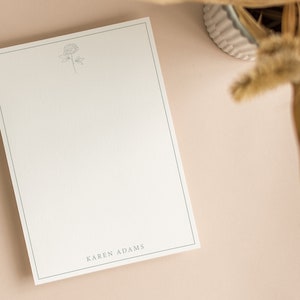 May include: A cream-colored notepad with a delicate floral illustration and a thin blue border. The name "KAREN ADAMS" is printed at the bottom. The notepad is on a light pink surface, with dried plants in the background.