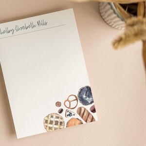 May include: A white notepad with the name "Shelby Elizabeth Miller" written at the top. The bottom right corner of the notepad features a watercolor illustration of various baked goods, including a pie, bread, and pretzels.
