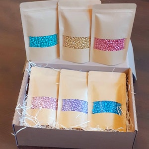 May include: A set of six small bags filled with colourful sprinkles. The bags are arranged in a box with shredded paper. The sprinkles are in shades of pink, blue, and gold.