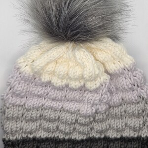 May include: A knitted hat with a fluffy grey pom pom. The hat is striped with white, light grey, dark grey, and black.