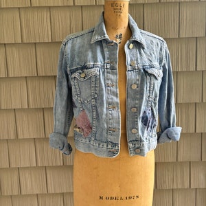 May include: A light blue denim jacket with patches on the front and sleeves. The jacket is on a dress form.