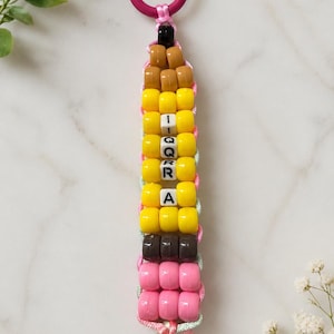 May include: A keyring featuring a pencil-shaped design made of colourful plastic pony beads. The beads are arranged in rows of tan, yellow, black, and pink. White letter beads spell out the name IQRA vertically in the centre of the yellow section.