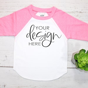 May include: A white and pink baseball tee shirt with the text "YOUR design HERE" printed on the front. The shirt is laying flat on a white wooden surface.