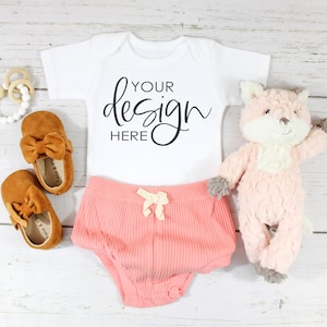 May include: A white baby onesie with the text "YOUR design HERE" in black script font. The onesie is paired with a pink ribbed baby bloomer and brown suede baby shoes with bows. A pink plush fox toy is also included.