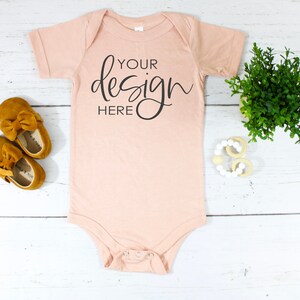 Pode incluir: Um body de bebê cor pêssego com mangas curtas e fecho de pressão. O body tem o texto "YOUR design HERE" impresso na frente em preto.