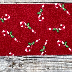 K&ouml;nnte beinhalten: Roter Stoff mit einem sich wiederholenden Muster aus wei&szlig;en Zuckerstangen und gr&uuml;nen Stechpalmenbl&auml;ttern.