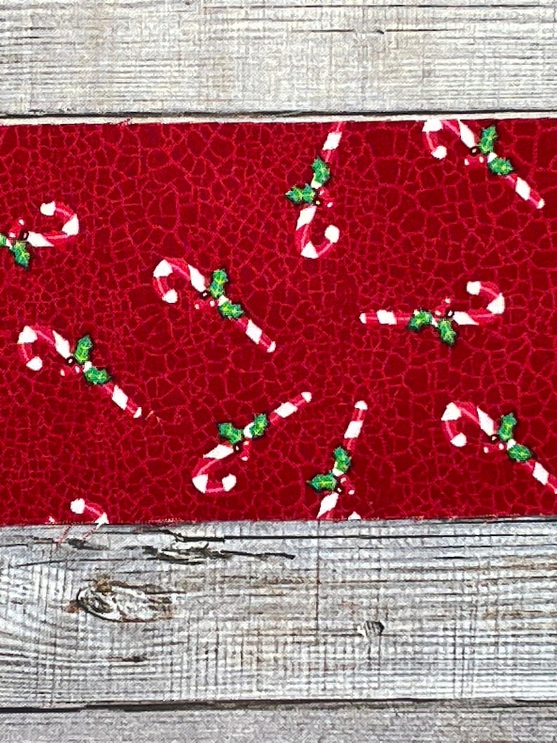 K&ouml;nnte beinhalten: Roter Stoff mit einem sich wiederholenden Muster aus wei&szlig;en Zuckerstangen und gr&uuml;nen Stechpalmenbl&auml;ttern.