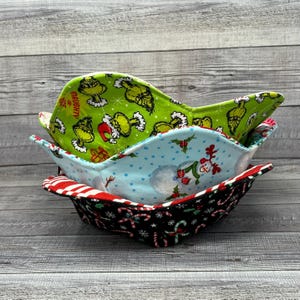 May include: Three fabric bowls stacked on top of each other. The top bowl is green with a Grinch pattern, the middle bowl is blue with a snowman pattern, and the bottom bowl is black with a candy cane pattern.
