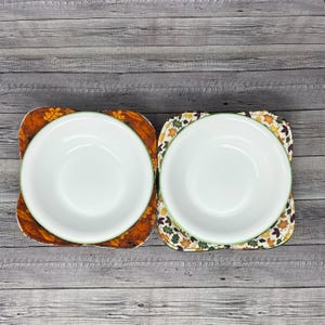 May include: Two white bowls with green rims are sitting on top of fabric placemats with a fall leaf pattern. The placemats are brown, orange, and yellow.