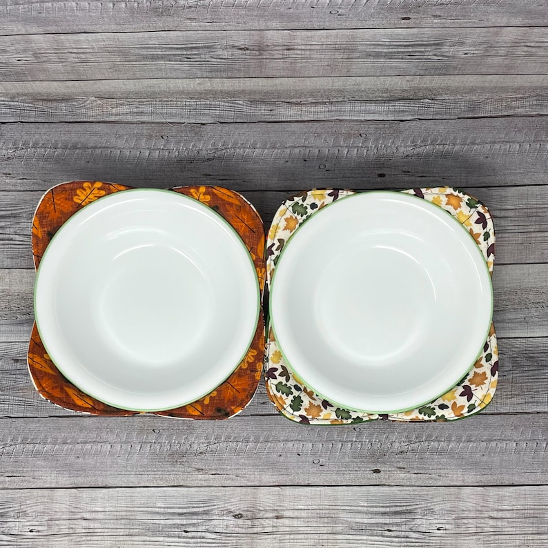 May include: Two white bowls with green rims are sitting on top of fabric placemats with a fall leaf pattern. The placemats are brown, orange, and yellow.