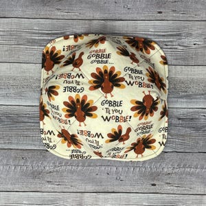 May include: A white fabric bowl cover with a Thanksgiving turkey pattern. The pattern features turkeys with the text "Gobble 'til you Wobble!" repeated throughout.