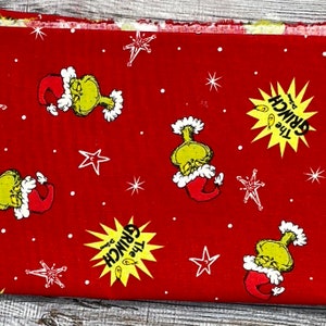 K&ouml;nnte beinhalten: Roter Stoff mit einem sich wiederholenden Muster aus dem Grinch mit einem Weihnachtsmann-Hut, einem gelben Stern mit dem Text "The Grinch" und wei&szlig;en Sternen.