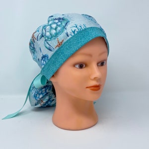 May include: A light blue scrub cap featuring a sea turtle pattern with teal accents. The cap has a teal band and a matching ribbon tie. The design includes sea turtles, starfish, and coral.