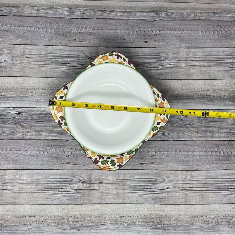 May include: A white ceramic bowl with a green and brown leaf print on the rim, sitting on a square fabric hot pad with a similar leaf print. The bowl is 9 inches in diameter.